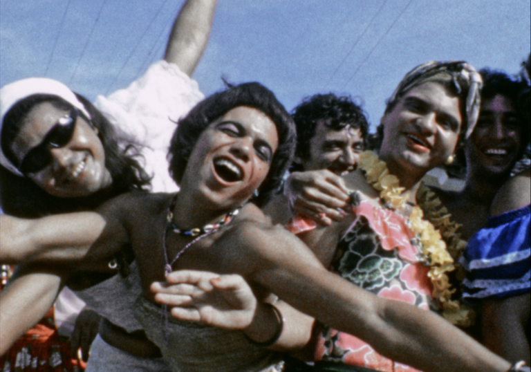 A group of queer people hold each other joyously in the sun. 