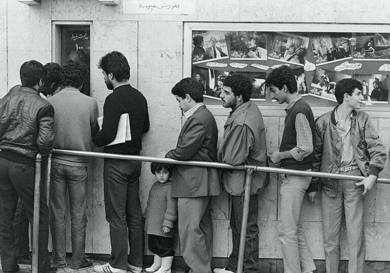 A black and white photograph of a group of men queuing up outside of an old movie theatre. 