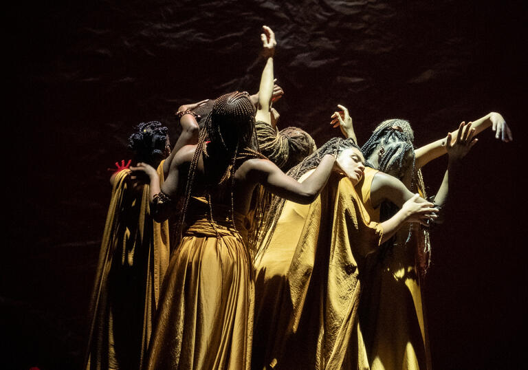 Members of the cast of The Golden Stool in long golden dresses, standing in a close huddle, some with their arms outstretched