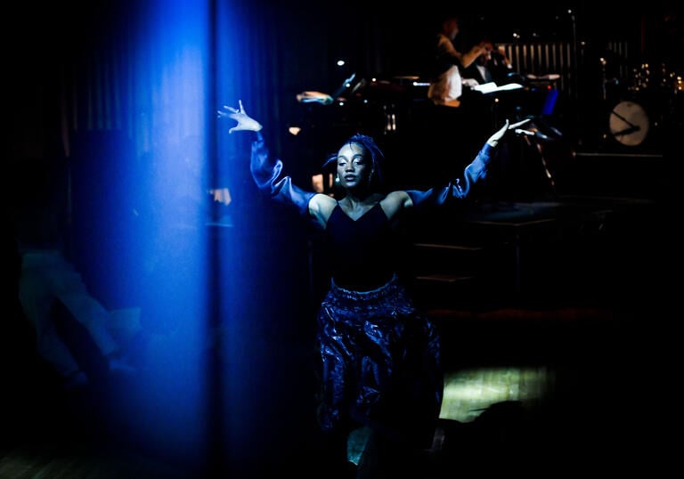 A person wearing black and blue walks with their hands in the air across the stage as a live band plays behind them.