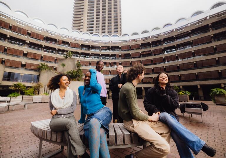 A group of six young people sat and standing, laughing in the Sculpture Court at the Barbican