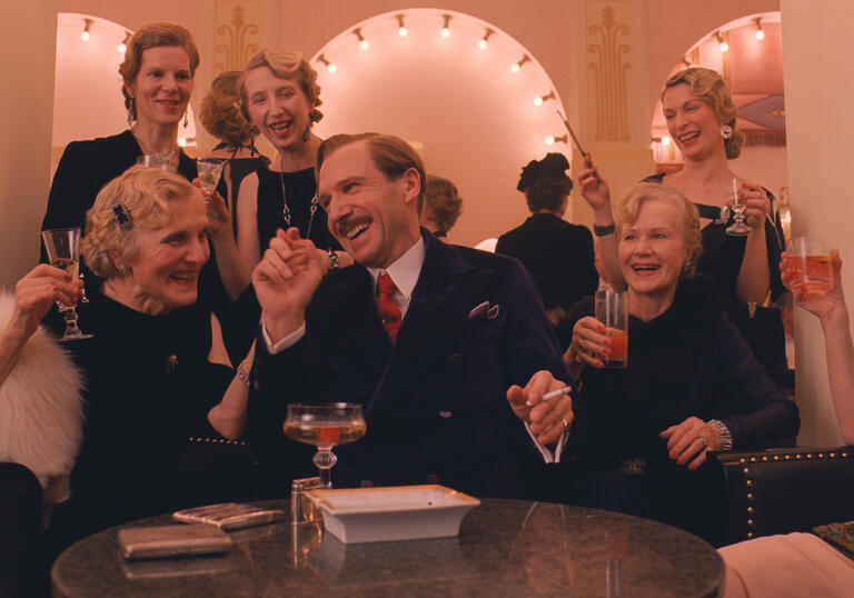 A hotelier sits smoking and laughing, surrounded by glamorous older women, in a nice looking hotel salon.