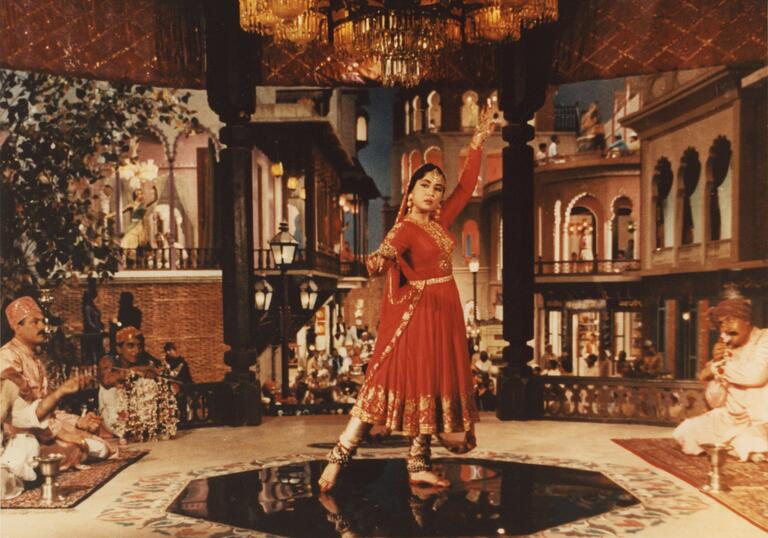 A woman in traditional Indian dress poses in a wonderfully decorated room, whilst men watch on.
