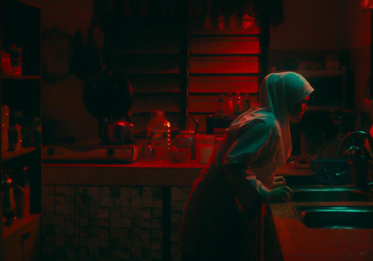 A young girl in a headscarf grasps onto the kitchen sink in a dark red kitchen at night.