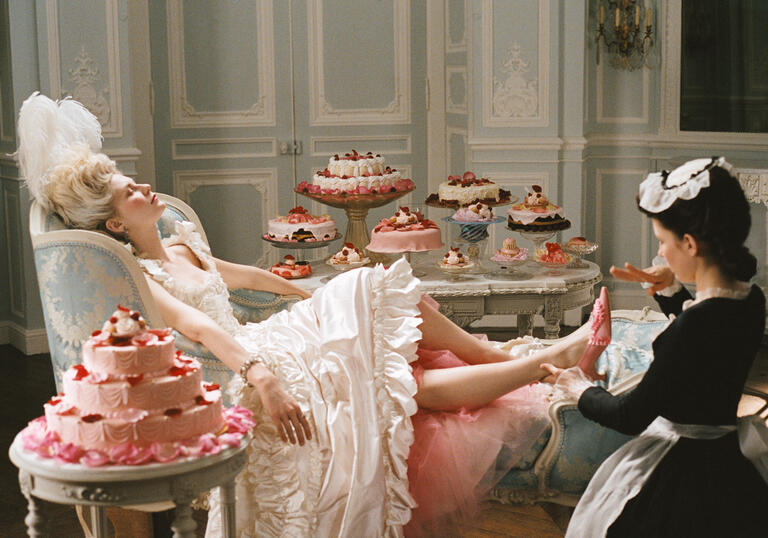 A queen lies back in a blue armchair, surrounded by decadent cakes, getting her feet rubbed by a maid.