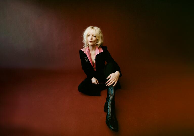 Jessica Pratt sits on dark red floor wearing a black suit and a pink shirt with large collar 