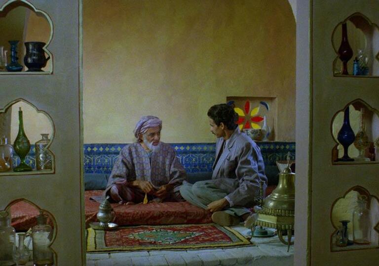 A young and an old man sit in a temple conversing, surrounded by lamps and glass jars.