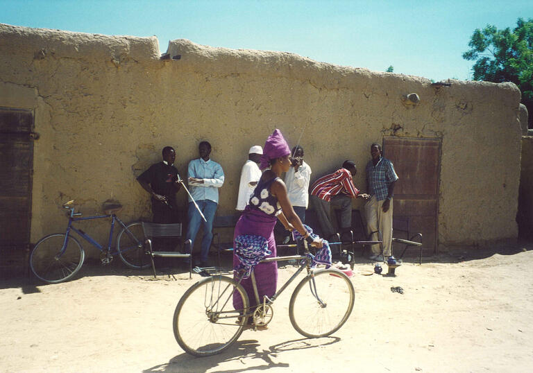 A woman passes a group of men on a bicycle in a desert village. 