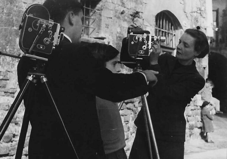 Margaret Tait stands by two large, old fashioned cameras with a man next to her in an image from the 1970s.