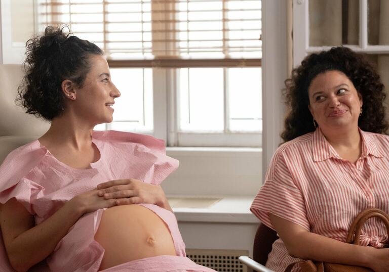 Two women wearing pink smile at each other, one has her stomach showing to reveal a large baby bump.