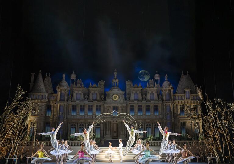 A group of dancers pose on a stage in front of a mansion, they are all dancing.