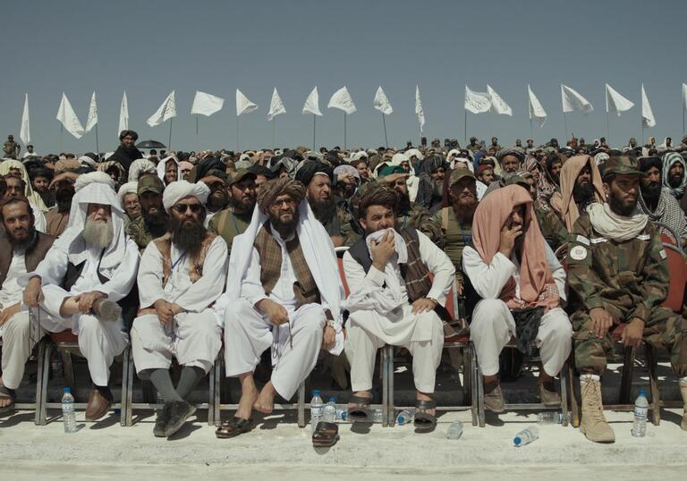 A group of men sit in a large outdoor seated area, in white desert robes.