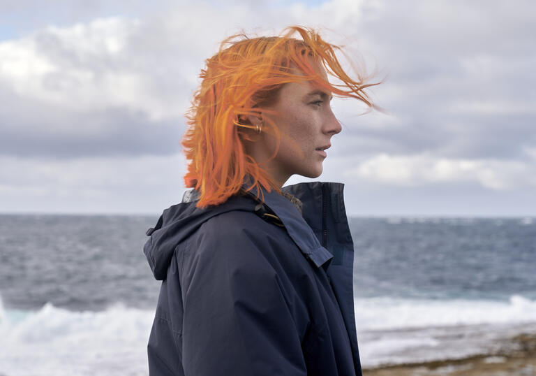 A woman with orange hair stands in front of a stormy sea. 