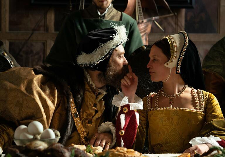 A man and a woman in tudor dress gaze into each other's eyes at a banquet, as the woman touches the man's nose.