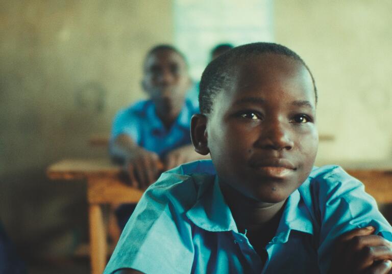 A child in a blue shirt is sat in a classroom looking up at the front the room, surrounded by classmates.