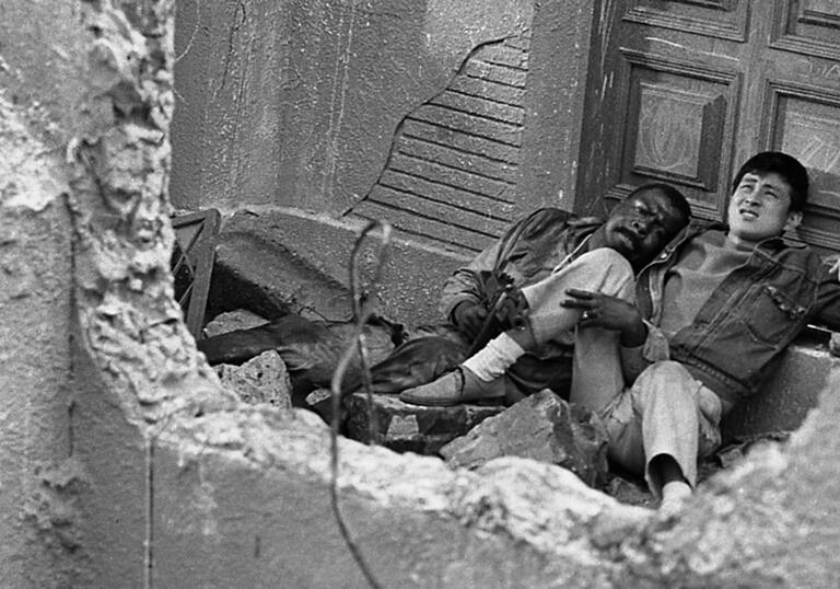 Two men lie by a door by a destroyed building. 