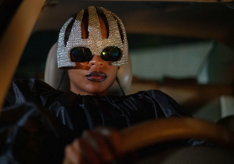 A woman with sunglasses, a sparkly helmet and red lipstick drives a car. 