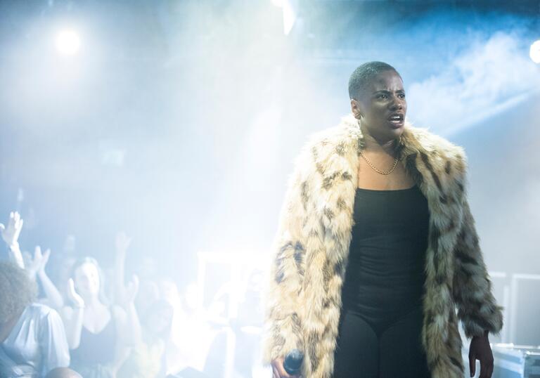 A person in a fur coat stands on stage as people dance in the background in a dark room lit by haze.