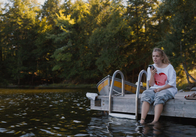 A young woman sits on the dock of a lake, her legs in the water.