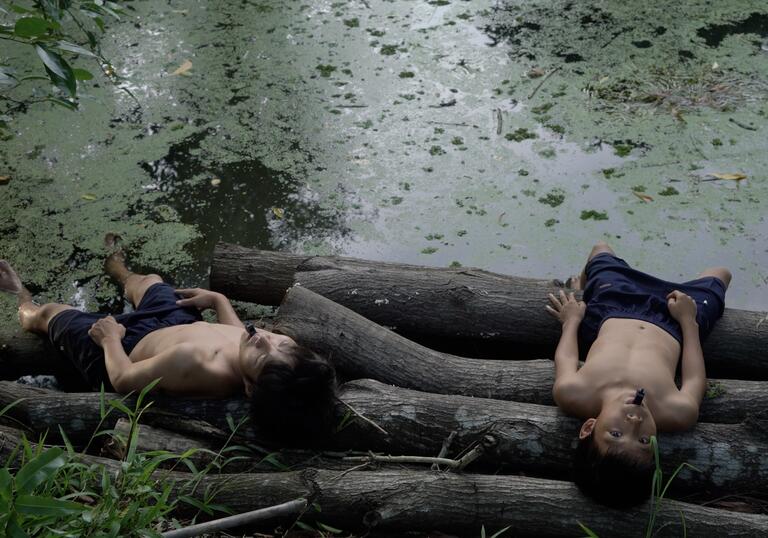 Two young boys lie on logs by a river covered in green algae. 