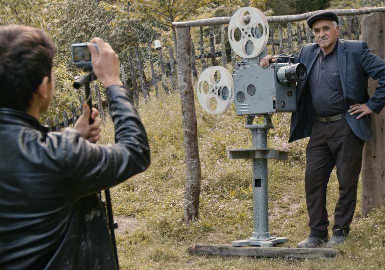 A man takes a photograph of another man standing next to an old cinema projector in a garden.