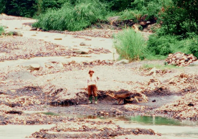 A young person sits on a rock in front of a wide, calm river.