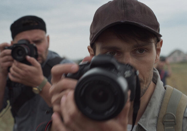 A young man with a professional camera looks ahead, whilst another man with a camera stands behind him, ready to shoot. 