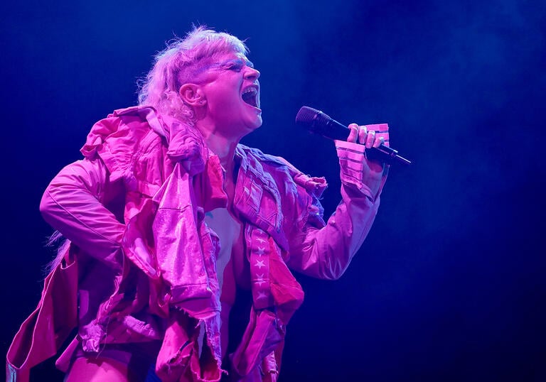 Peaches in pink stage lighting sings while holding a microphone 