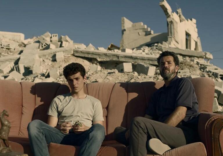 Two men sit on a couch outdoors, surrounded by rumble