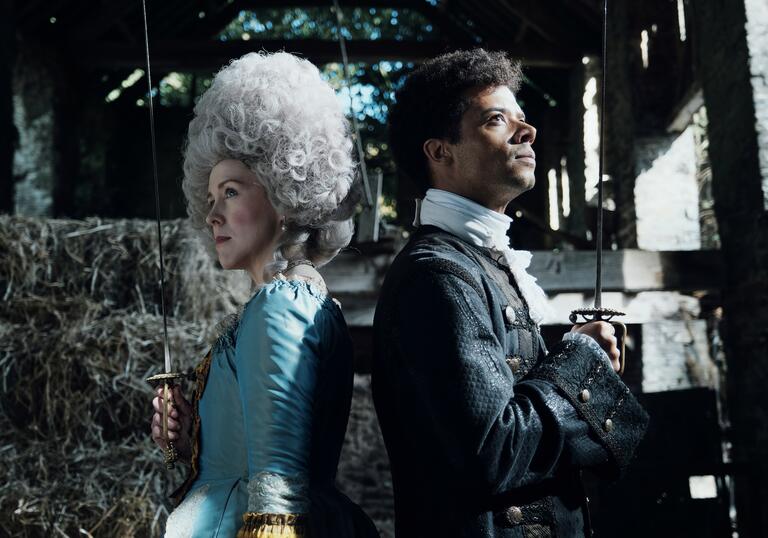 A woman and man in 18th-century clothing and wigs stand back-to-back, with both holding swords pointed skyward.