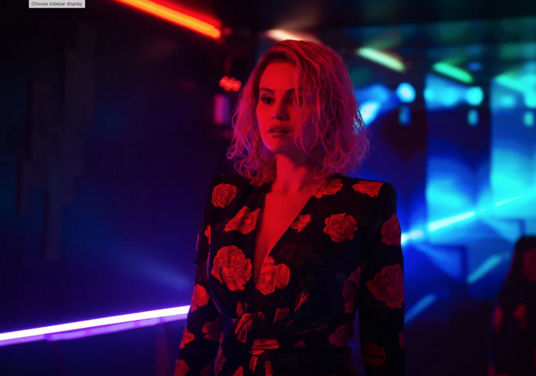 A woman stands in a blue, dark corridor, bathed in red light. 