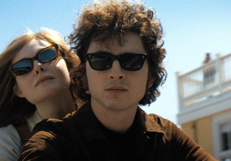 Two people wearing sunglasses ride through an American town with a blue sky overhead.