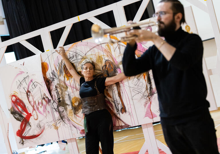 Rachel, a white woman with short cropped hair, wearing black, and Freddie, a white male, with a full beard, wearing a black long sleeved top with a black skirt, in rehearsals for PostHuman. Freddie is out of focus in the foreground playing a silver trumpet, and Rachel is in focus leaning back on a large canva with arms out and eyes closed.