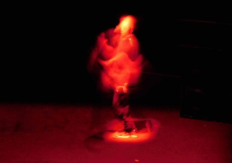 A person moves through a dark space, they are lit by a red light so that you can't see them as much as just a red figure moving through the space.