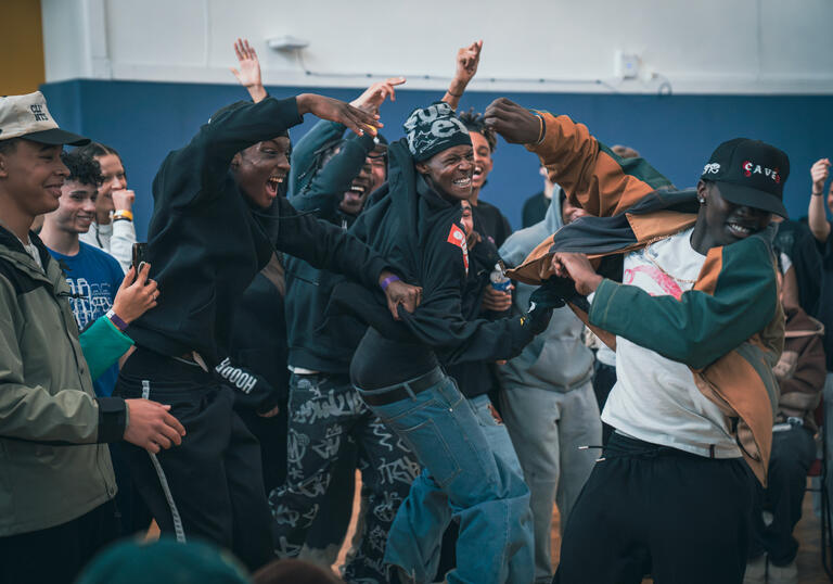 A group of hip hop dancers cheer and embrace in a rehearsal room.