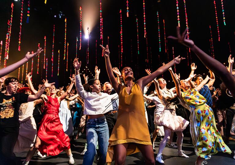 A group of dancers dressed in colourful clothing move on stage with their arms in the air.