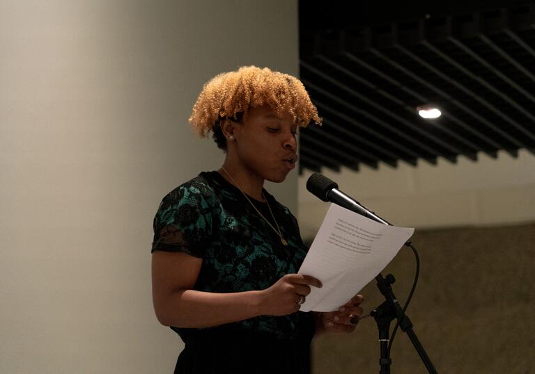 A Barbican Young Poet holds a piece of paper and reads into a microphone. 