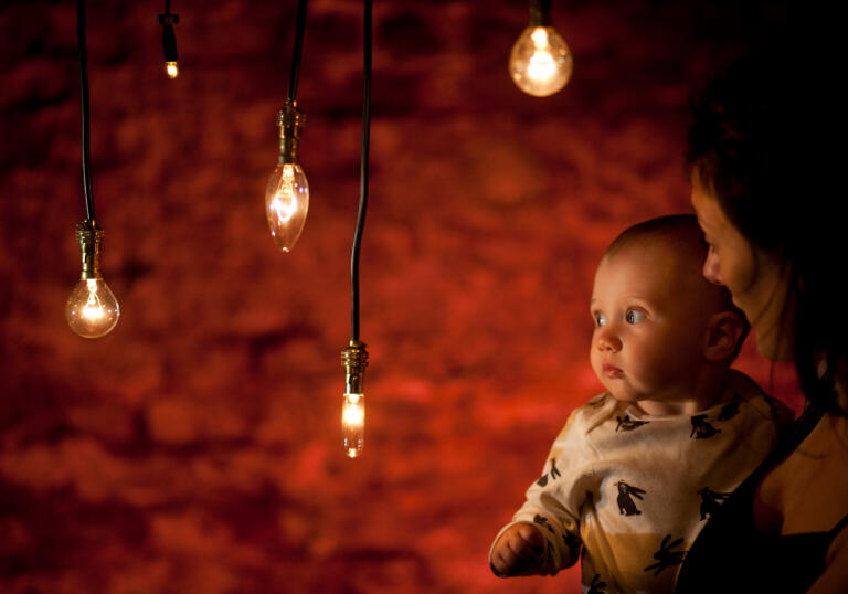 A person holding a baby in a dark room staring at lightbulbs hanging from the ceiling