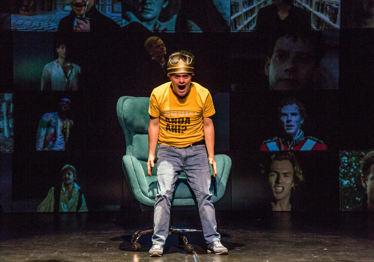 A person in a yellow shirt and blue jeans standing over a green sofa chair, shouting. They are against a background of screens with each screen showing pictures of different men.