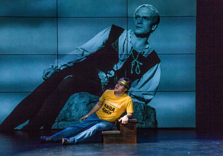 A person in a yellow shirt and blue jeans laying down on their side on a pair of stairs. They are against a background of screen which is showing a picture of another person dressed in historical clothing, also laying on their side over a rock.  