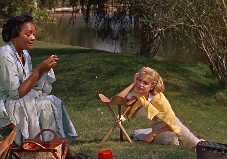 An older woman sits on the grass with a younger woman at her feet.