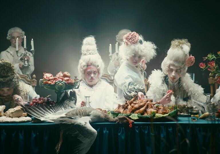 A group of actors in wigs sit at a table covered in food. 