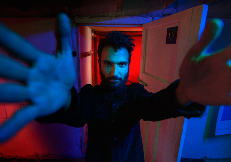 A man with hark hair and beard with his hands extended forward reaching for the camera