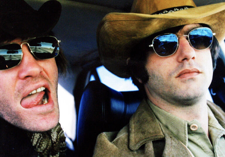 Two men sit in a vehicle wearing sunglasses. 