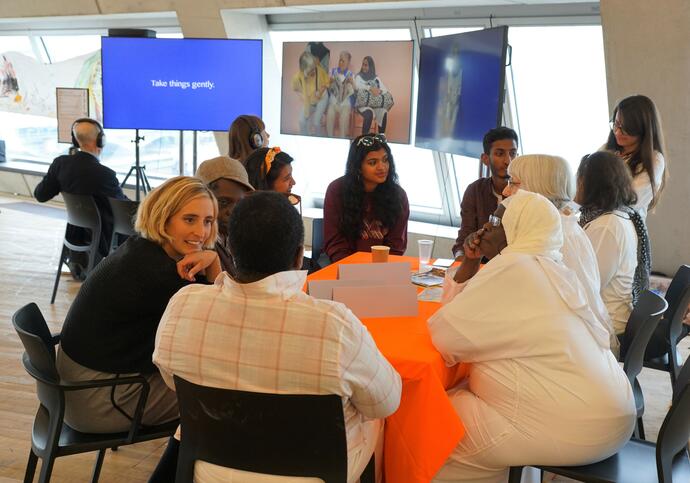 Group of individuals huddled in a circle smiling and talking. There is a projector screen behind them with a blue screen.