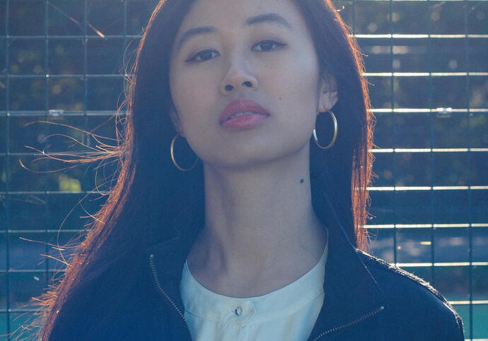 image of poet Christy who is standing outside with the sun glaring into the camera. Christy has long black hair and is wearing a black jacket with white tshirt.