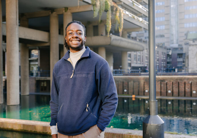 Image of Jordan standing in front of Barbican lake, wearing blue zip up fleece