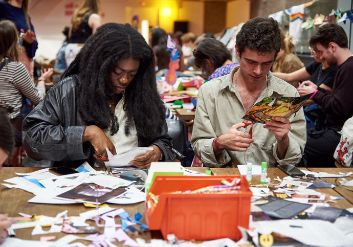 Two people sat at table making a collage, one in black leather jacket and the other in a beige shirt