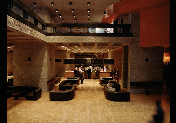 Foyer space with curved brown sofas and people at the end of the room standing at a desk, from the Barbican Level 1 foyer in 1982