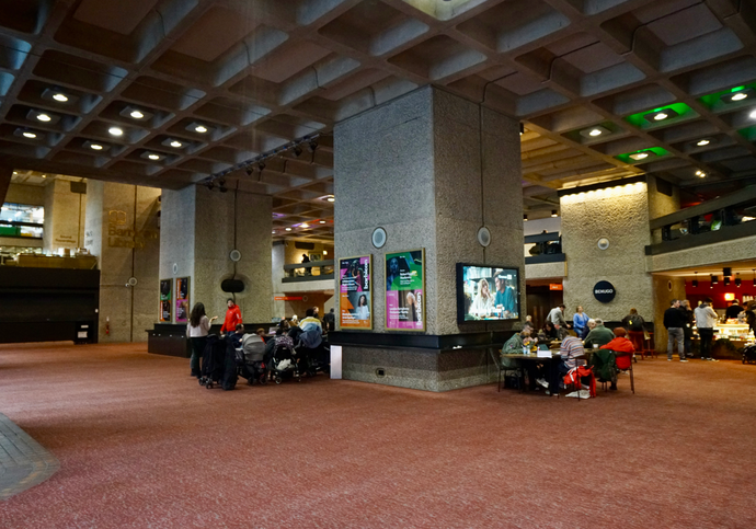 The Barbican Level G foyer, with patchy lighting and some people sitting and gathering in the space